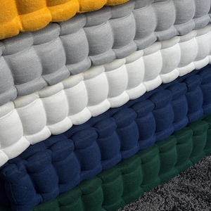Peut inclure: Gros plan sur une pile de coussins touffet&eacute;s color&eacute;s. Les coussins sont dans les tons de jaune, gris, blanc, bleu marine et vert. Le tissu a un aspect textur&eacute; et matelass&eacute;, avec un motif r&eacute;p&eacute;titif de carr&eacute;s arrondis.