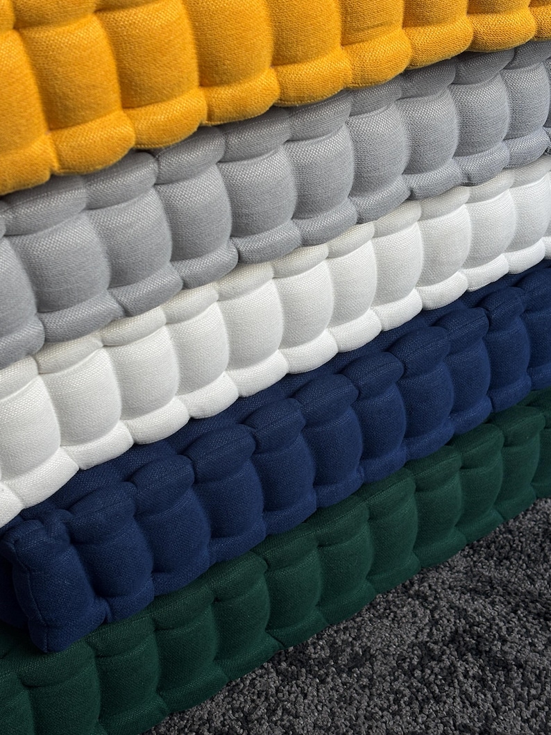 Peut inclure: Gros plan sur une pile de coussins touffet&eacute;s color&eacute;s. Les coussins sont dans les tons de jaune, gris, blanc, bleu marine et vert. Le tissu a un aspect textur&eacute; et matelass&eacute;, avec un motif r&eacute;p&eacute;titif de carr&eacute;s arrondis.