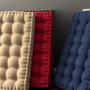 Three thick, tufted floor cushions leaning against a gray wall, arranged side by side on the floor; cream-colored on the left, red in the center, and navy blue on the right.