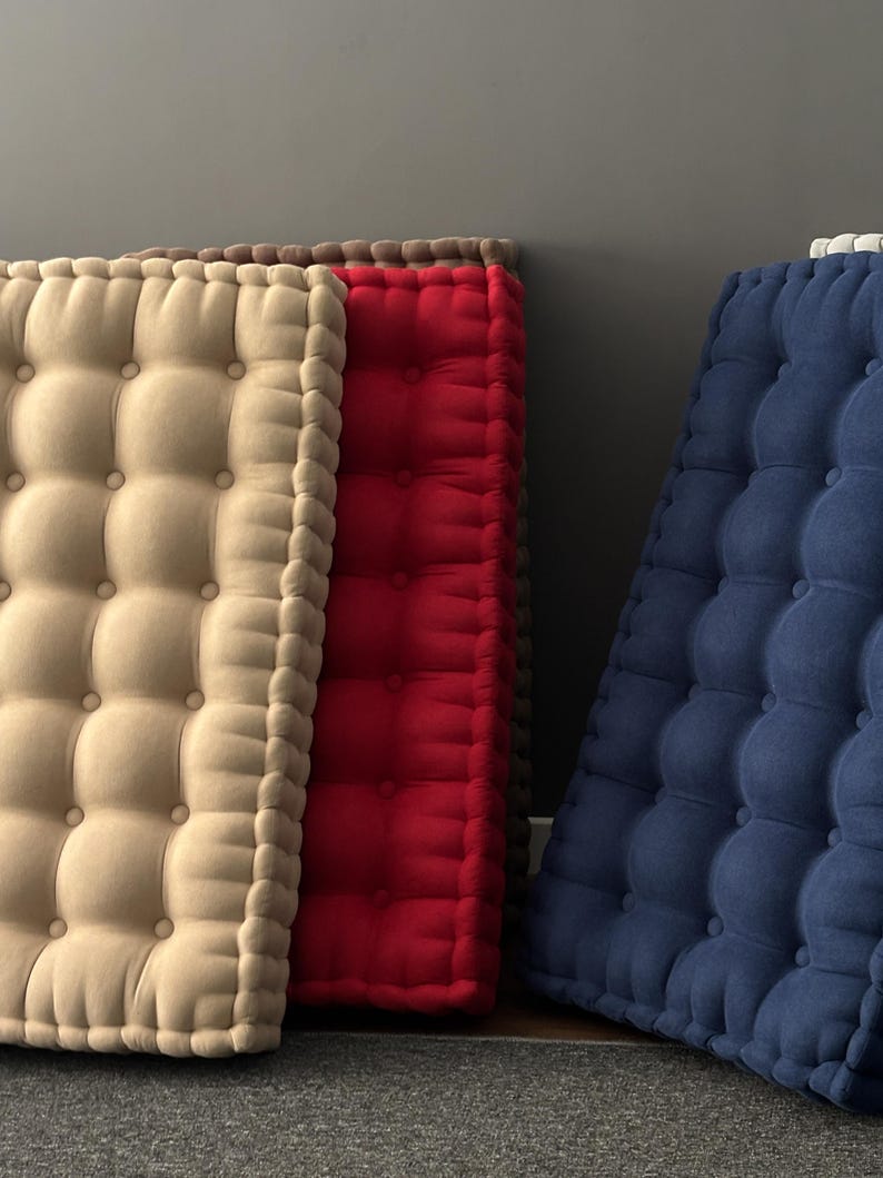 Three thick, tufted floor cushions leaning against a gray wall, arranged side by side on the floor; cream-colored on the left, red in the center, and navy blue on the right.