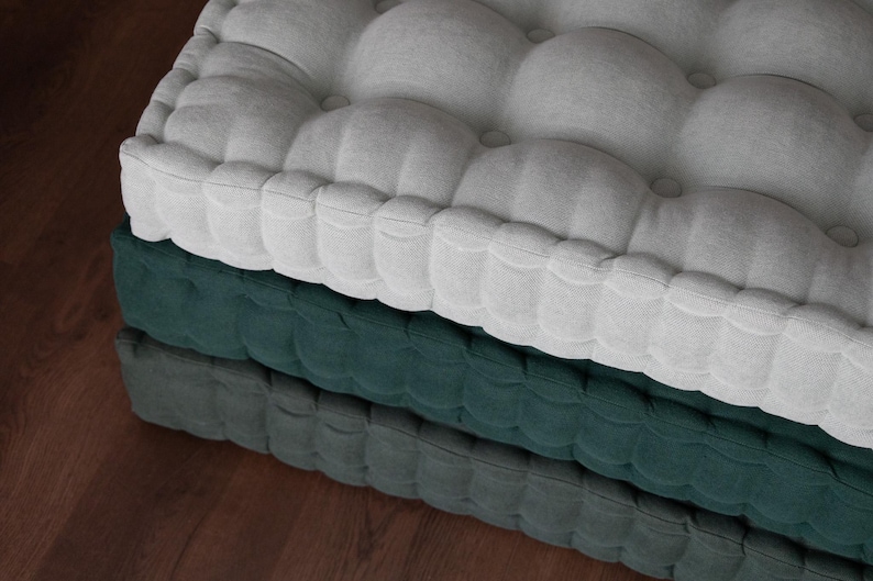 Close-up of stacked tufted floor cushions on a wooden floor, with a light gray cushion on top, a deep green cushion in the middle, and a darker green cushion on the bottom, showing soft fabric texture and button detailing.