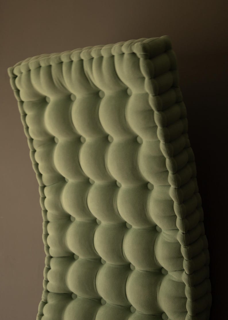 Side view of a thick, square tufted cushion in soft olive green fabric, showing deep button detailing and rounded edges against a neutral wall.