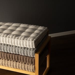 Square, thick tufted cushions stacked on a wooden bench, with a light gray cushion on top, a medium gray cushion in the middle, and a taupe cushion on the bottom, set against a dark gray wall.