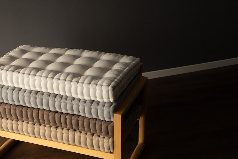Square, thick tufted cushions stacked on a wooden bench, with a light gray cushion on top, a medium gray cushion in the middle, and a taupe cushion on the bottom, set against a dark gray wall.