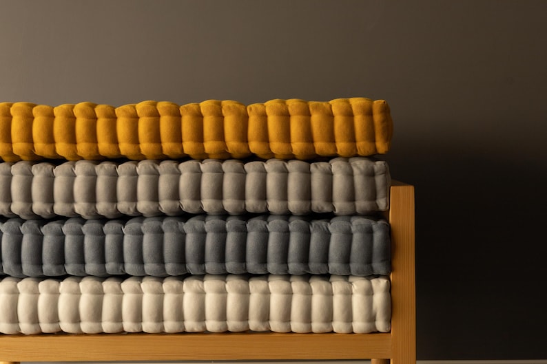 A wooden bench holding four neatly stacked, thick tufted cushions in warm mustard yellow, light gray, dark gray, and off-white, set against a smooth gray wall.