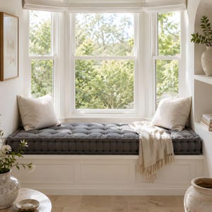 A gray tufted floor cushion fully covers a bright bay window seating area decorated in white and stone tones. Cream pillows, a light fringed throw, ceramic vases, and natural daylight create an airy, minimalist home decor atmosphere.