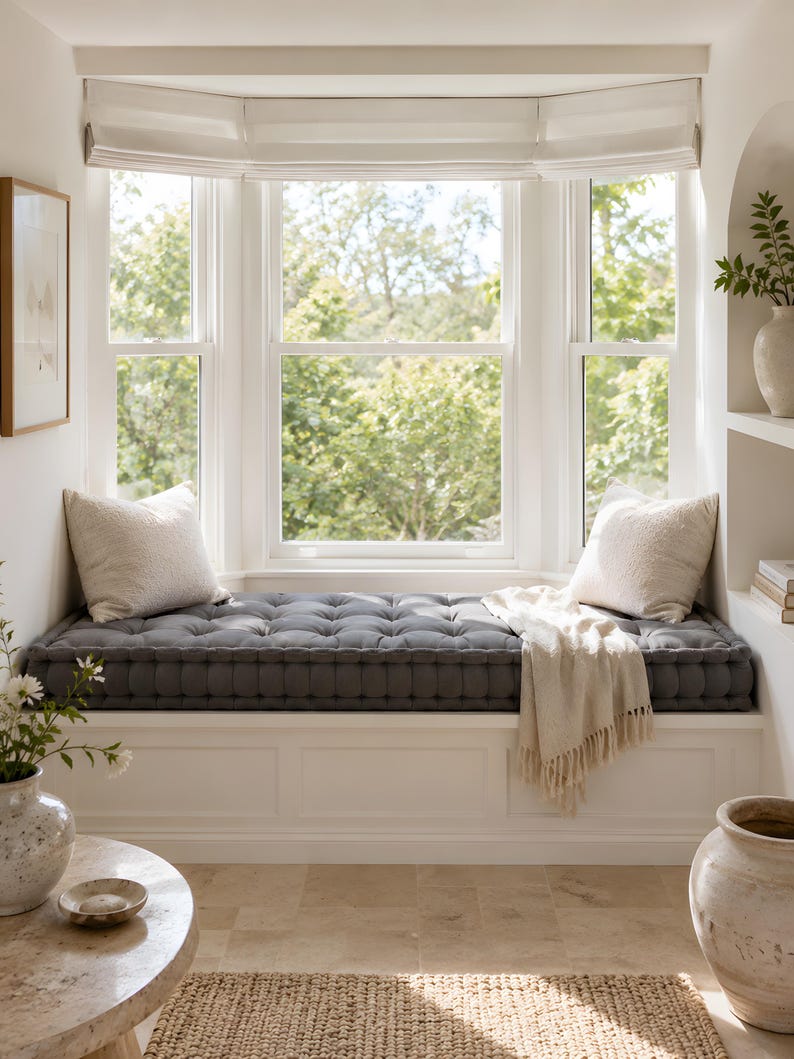 A gray tufted floor cushion fully covers a bright bay window seating area decorated in white and stone tones. Cream pillows, a light fringed throw, ceramic vases, and natural daylight create an airy, minimalist home decor atmosphere.