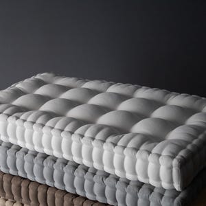 Square, thick tufted cushions stacked on a wooden bench, with a light gray cushion on top, a medium gray cushion in the middle, and a taupe cushion on the bottom, set against a dark gray wall.