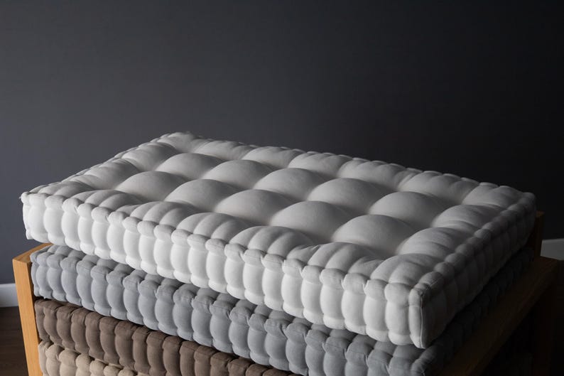 Square, thick tufted cushions stacked on a wooden bench, with a light gray cushion on top, a medium gray cushion in the middle, and a taupe cushion on the bottom, set against a dark gray wall.