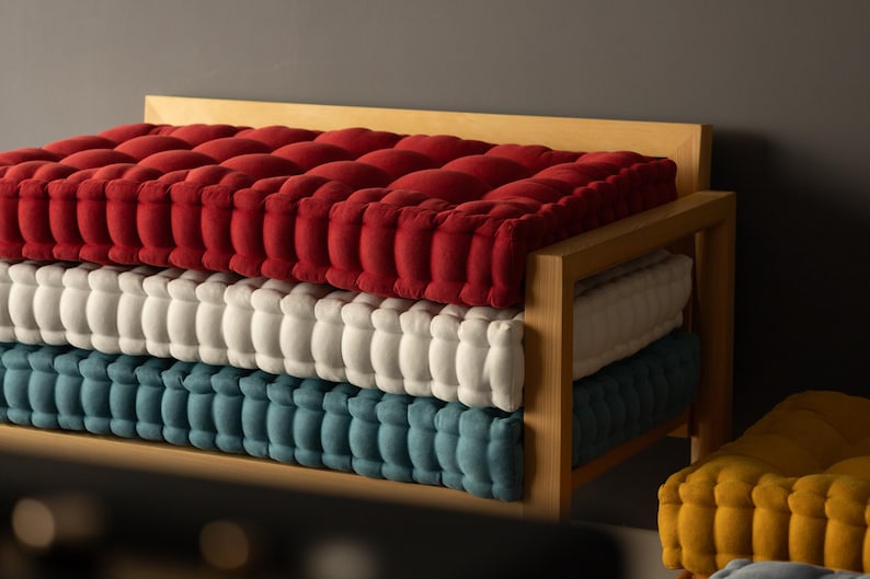 Window bench decoration ideas. A wooden bench with neatly stacked, thick tufted cushions in three layers: deep red on top, off-white in the middle, and muted teal on the bottom, set against a gray wall; a mustard-yellow tufted cushion is partially visible in the foreground.