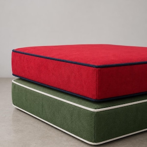 May include: A stack of two square floor cushions. The top cushion is bright red with dark blue piping along the edges. The bottom cushion is forest green with white piping along the edges. Both cushions feature a textured fabric finish.