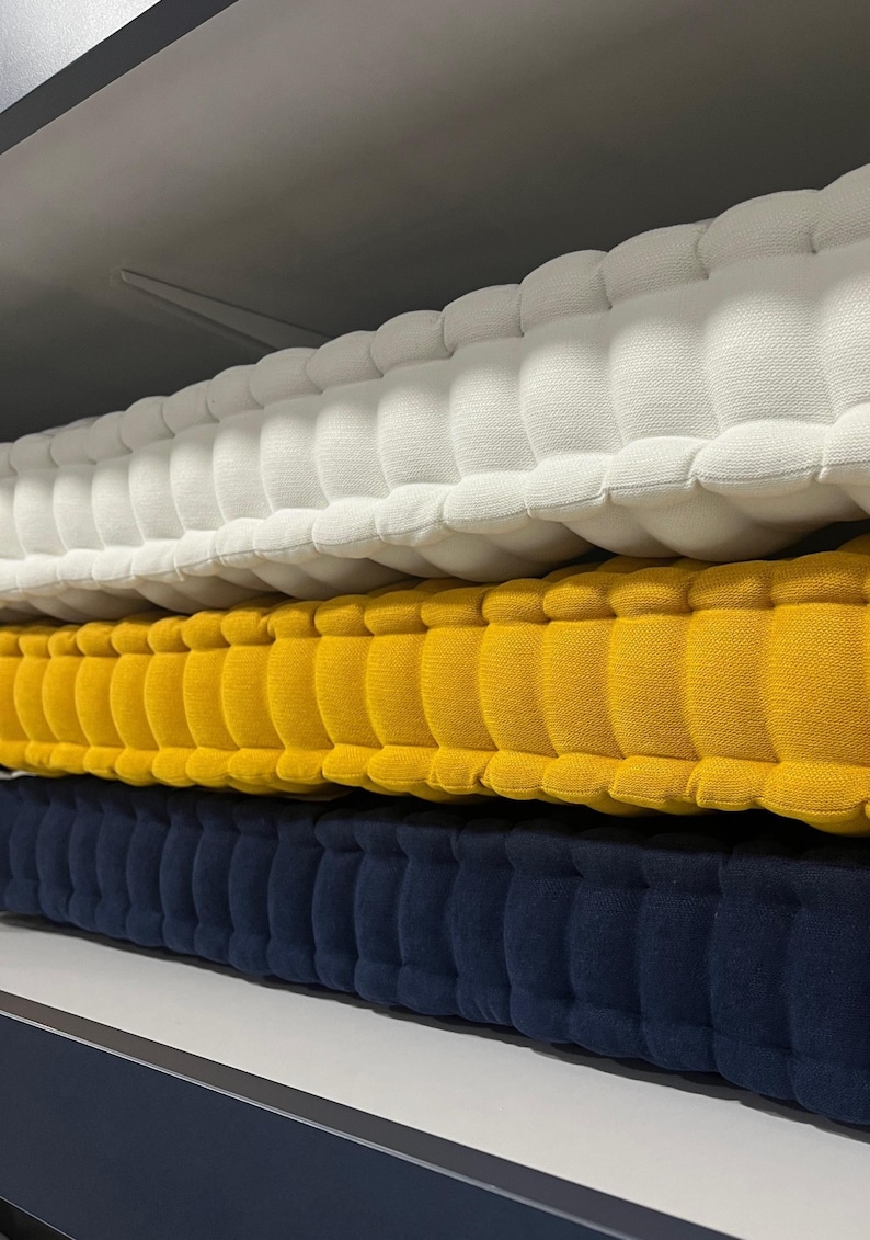 Stacked tufted cushions on a shelf, shown in close-up from the side, with textured fabric layers in off-white on top, mustard yellow in the middle, and navy blue on the bottom.