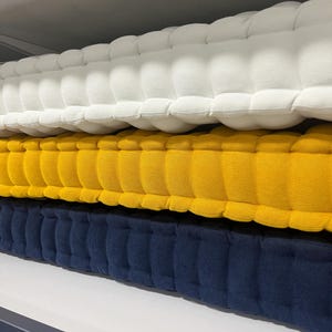 Stacked tufted cushions on a shelf, shown in close-up from the side, with textured fabric layers in off-white on top, mustard yellow in the middle, and navy blue on the bottom.
