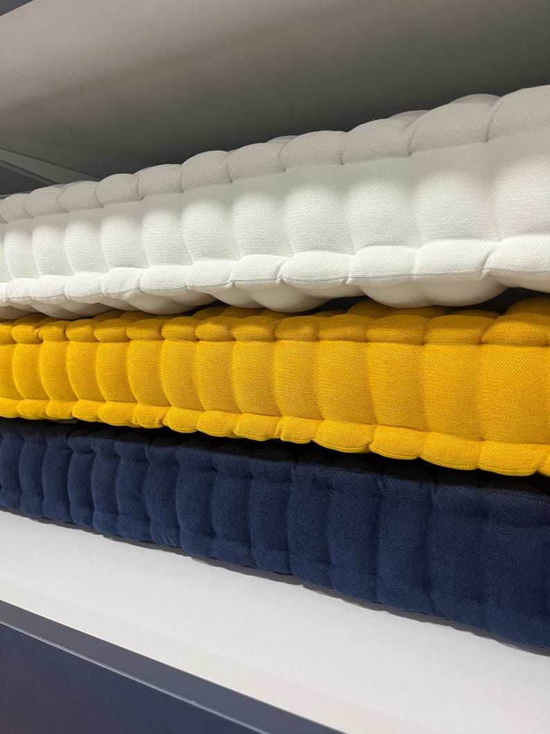 Stacked tufted cushions on a shelf, shown in close-up from the side, with textured fabric layers in off-white on top, mustard yellow in the middle, and navy blue on the bottom.