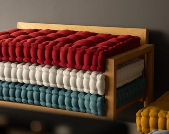 Puede incluir: Un marco de banco de madera clara que sostiene una pila de tres cojines de suelo rectangulares capitoné en rojo, blanco y azul cerceta. Un cojín capitoné amarillo separado se coloca a la derecha del banco, mostrando un patrón de rejilla.