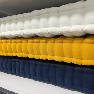 Peut inclure: Trois coussins touffet&eacute;s, blanc, jaune et bleu marine, empil&eacute;s sur une &eacute;tag&egrave;re. Les coussins ont un aspect matelass&eacute; textur&eacute;, avec une s&eacute;rie d'indentations arrondies et espac&eacute;es uniform&eacute;ment. Les couleurs sont unies et vibrantes, cr&eacute;ant un affichage visuellement attrayant.