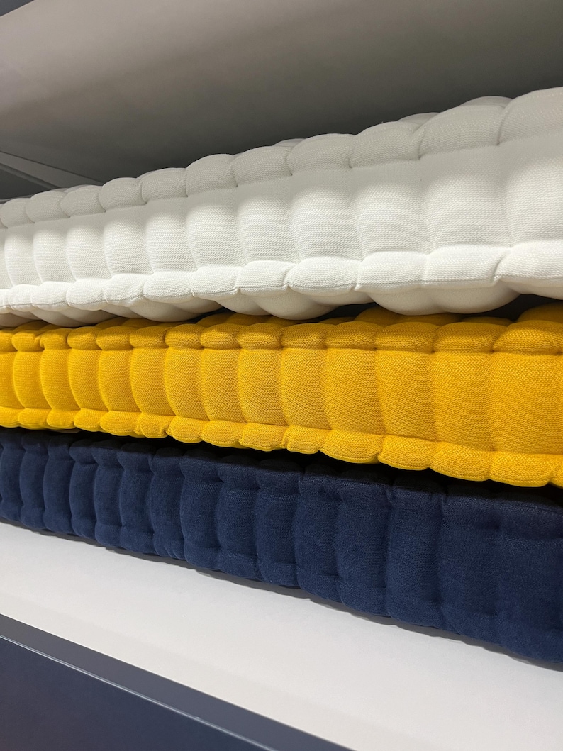 Peut inclure: Trois coussins touffet&eacute;s, blanc, jaune et bleu marine, empil&eacute;s sur une &eacute;tag&egrave;re. Les coussins ont un aspect matelass&eacute; textur&eacute;, avec une s&eacute;rie d'indentations arrondies et espac&eacute;es uniform&eacute;ment. Les couleurs sont unies et vibrantes, cr&eacute;ant un affichage visuellement attrayant.
