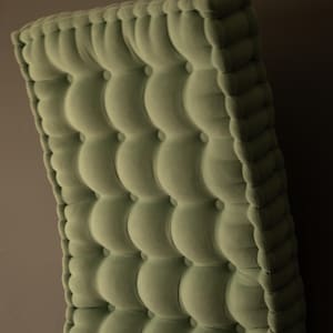 Side view of a thick, square tufted cushion in soft olive green fabric, showing deep button detailing and rounded edges against a neutral wall.