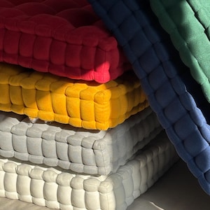 May include: A stack of square, tufted cushions in various colors. The colors include red, yellow, gray, and white, with additional cushions in navy blue and green. The cushions have a textured, grid-like pattern.