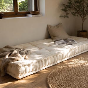May include: A cream-coloured floor mattress with a tufted design, a beige pillow, and a folded blanket. An open book rests on the mattress. A woven rug and a potted plant are also in the room.