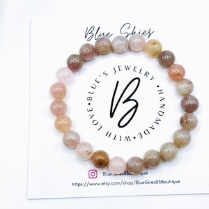 May include: A pink and brown gemstone bracelet with a white card behind it. The card has the text "Blue Skies Jewelry Handmade with Love Blue's" on it. The card also has a website address and a social media handle.