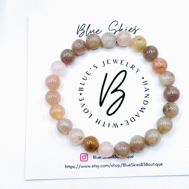 May include: A pink and brown gemstone bracelet with a white card behind it. The card has the text "Blue Skies Jewelry Handmade with Love Blue's" on it. The card also has a website address and a social media handle.