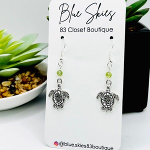 May include: Silver turtle earrings with green beads hanging from silver hooks. The earrings are displayed on a white card with the text "Blue Skies 83 Closet Boutique" and the Instagram handle "@blue.skies83boutique".