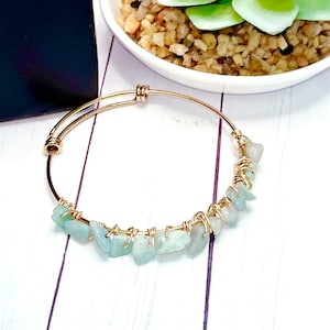 May include: Gold-coloured adjustable bangle bracelet with light blue-green gemstone accents. The gemstones are irregularly shaped and are secured with gold wire. The bracelet is displayed on a white wooden surface.