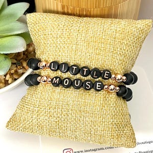 Op de afbeelding: Twee elastische armbanden met matte zwarte kralen en metallic roségoudkleurige tussenkralen. De bovenste armband toont het woord LITTLE en de onderste armband toont het woord MOUSE in witte letters op zwarte ronde kralen.