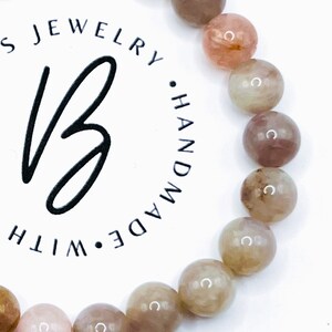 May include: A bracelet made with light pink, brown, and beige gemstone beads. The bracelet is handmade and features a logo with the text "JEWELRY &bull; HANDMADE &bull; WITH" in a circle.