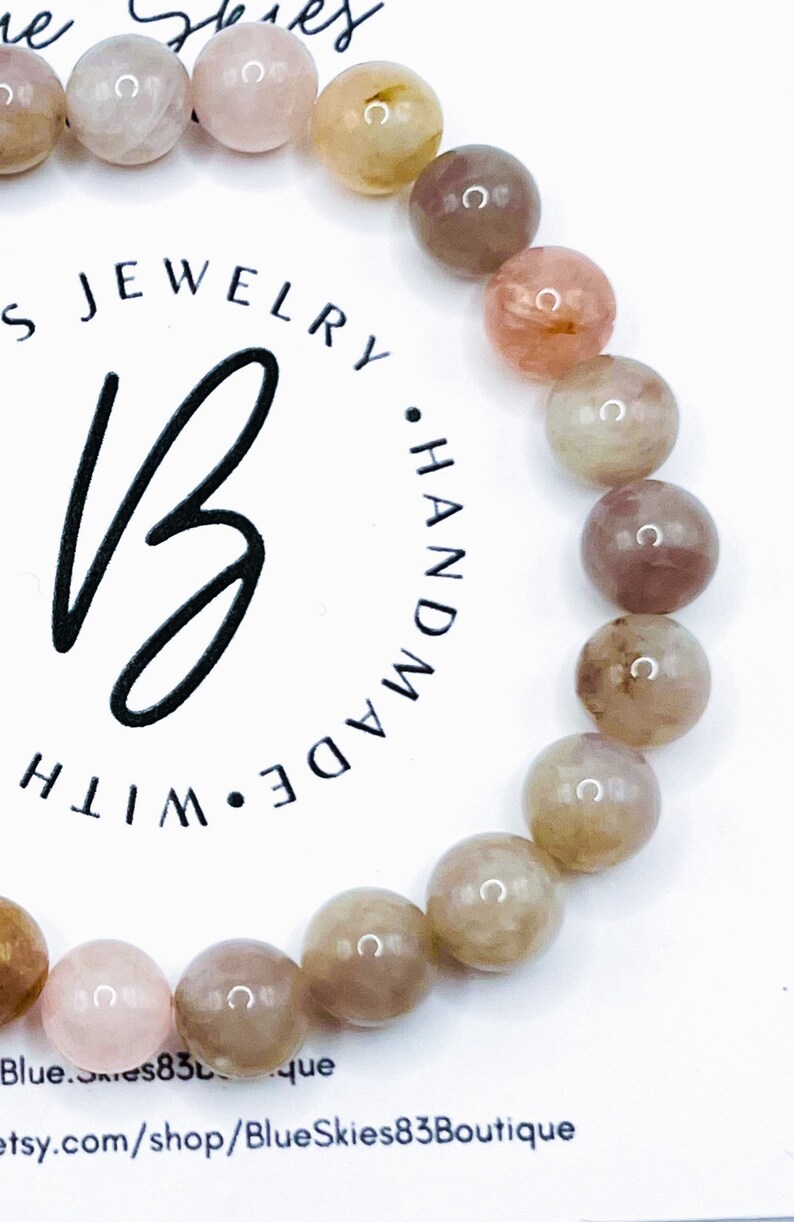 May include: A bracelet made with light pink, brown, and beige gemstone beads. The bracelet is handmade and features a logo with the text "JEWELRY &bull; HANDMADE &bull; WITH" in a circle.
