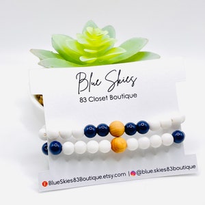 May include: Two bracelets with white and navy blue beads and a wooden bead. The bracelets are displayed on a white card with the text "Blue Skies 83 Closet Boutique".
