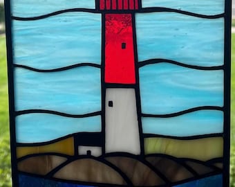 Può includere: Una finestra in vetro colorato che raffigura un faro rosso e bianco con un tetto giallo, che si erge su una costa rocciosa marrone con acqua blu in primo piano. Il cielo è blu con linee ondulate bianche.