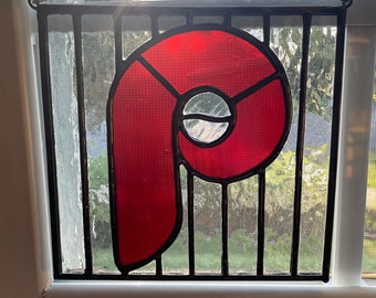 May include: A red stained glass letter 'P' hanging in a window. The letter is made of multiple pieces of glass and is framed in black metal.