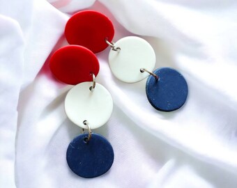 May include: Earrings featuring a series of red, white, and navy blue circular pendants. The pendants are connected by small silver hoops and hang against a white fabric background. The earrings have a modern, geometric design.
