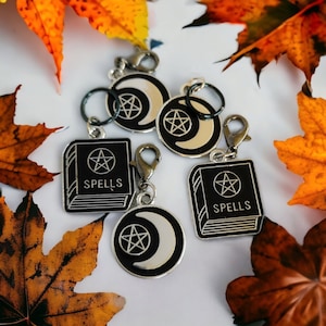 May include: Four silver charms with black enamel. Each charm features a crescent moon and pentagram design. Two charms have the word "Spells" written on them. The charms are on a white background with fall leaves.