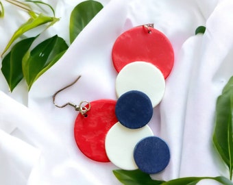 May include: A pair of handmade earrings featuring a red, white, and navy blue circular design. The earrings are composed of layered, flat circles, with a silver-colored hook. The earrings are displayed on a white fabric background with green leaves.