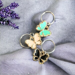 May include: Three butterfly-shaped keychains in pastel colors: light pink, mint green, and black. Each butterfly has a gold-colored outline and is attached to a silver-colored ring. The keychains are arranged on a gray fabric surface.