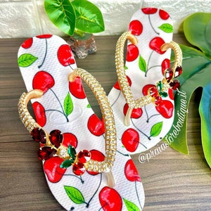 May include: White flip flops with a cherry print and gold straps adorned with rhinestones. The straps are decorated with red and green gems.