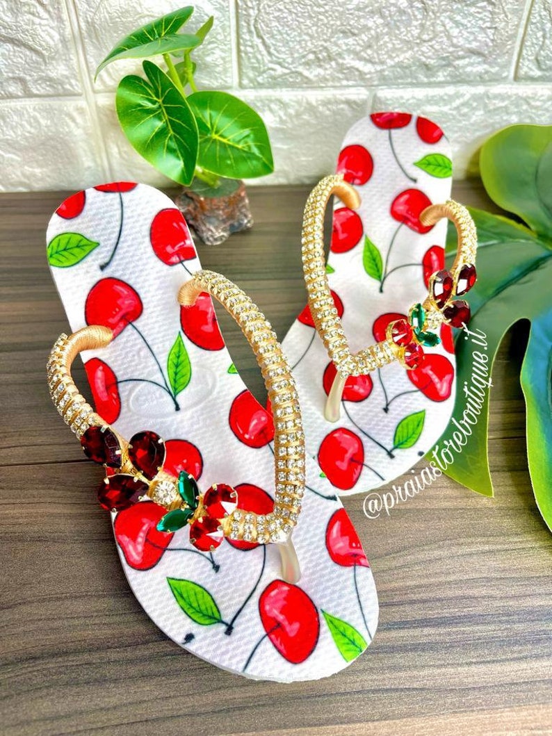 May include: White flip flops with a cherry print and gold straps adorned with rhinestones. The straps are decorated with red and green gems.