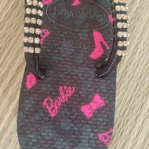 May include: Black flip-flop with a pink Barbie logo and pink bows. The flip-flop has a gold chain with a pink Barbie charm and rhinestones on the straps.
