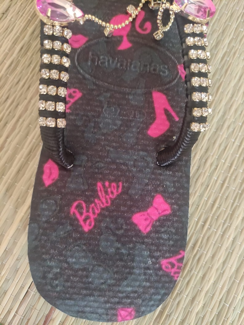 May include: Black flip-flop with a pink Barbie logo and pink bows. The flip-flop has a gold chain with a pink Barbie charm and rhinestones on the straps.