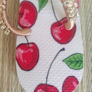 May include: White flip-flop with a red cherry print and a gold strap with rhinestones.