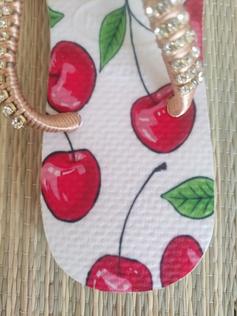May include: White flip-flop with a red cherry print and a gold strap with rhinestones.