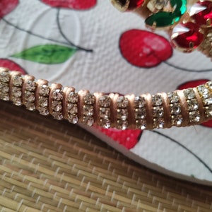 May include: Close-up of a gold-toned strap with rows of clear rhinestones. The strap is on a white background with a cherry pattern.