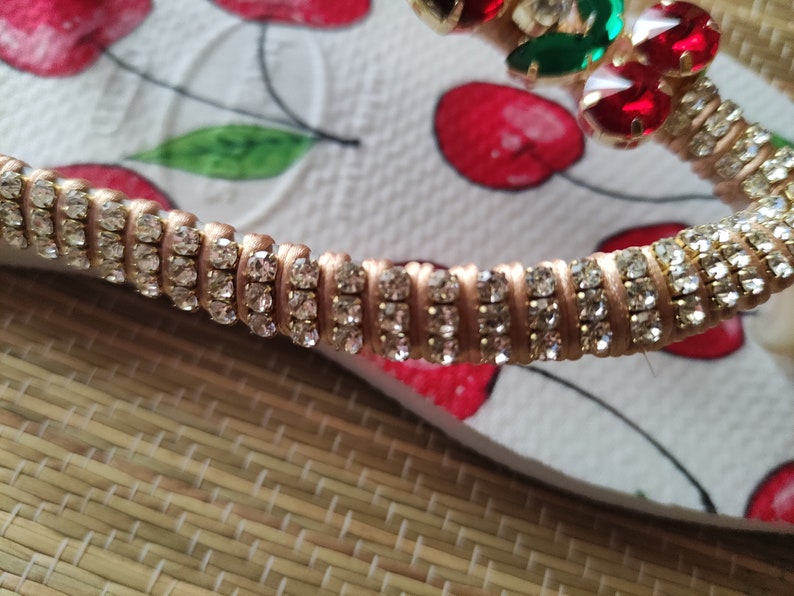May include: Close-up of a gold-toned strap with rows of clear rhinestones. The strap is on a white background with a cherry pattern.