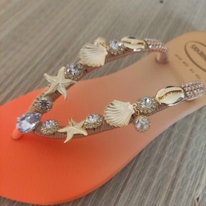 Puede incluir: Sandalia de playa naranja y melocot&oacute;n adornada con conchas marinas, estrellas de mar y brillantes pedrer&iacute;a. La sandalia presenta una correa dorada y est&aacute; adornada con varios dijes con tem&aacute;tica de playa.