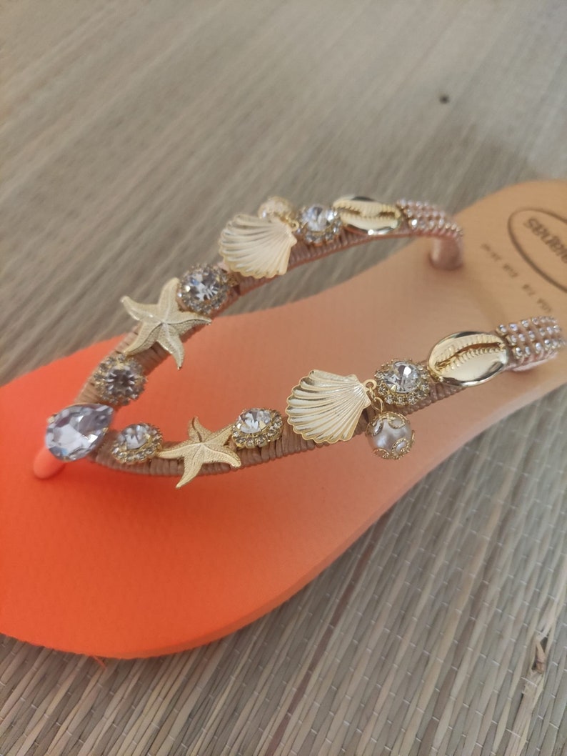Puede incluir: Sandalia de playa naranja y melocot&oacute;n adornada con conchas marinas, estrellas de mar y brillantes pedrer&iacute;a. La sandalia presenta una correa dorada y est&aacute; adornada con varios dijes con tem&aacute;tica de playa.
