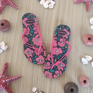 Puede incluir: Un par de chanclas rosas con estampado floral y el texto "havaianas" en las tiras. Las chanclas est&aacute;n sobre una superficie de madera con conchas y estrellas de mar.