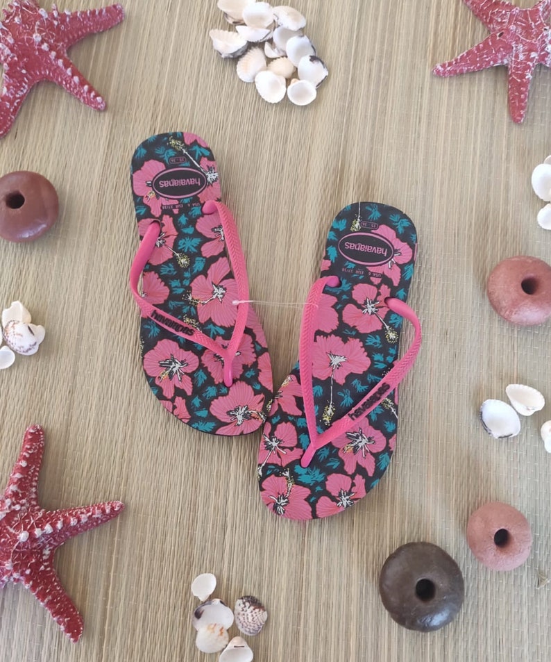Puede incluir: Un par de chanclas rosas con estampado floral y el texto "havaianas" en las tiras. Las chanclas est&aacute;n sobre una superficie de madera con conchas y estrellas de mar.